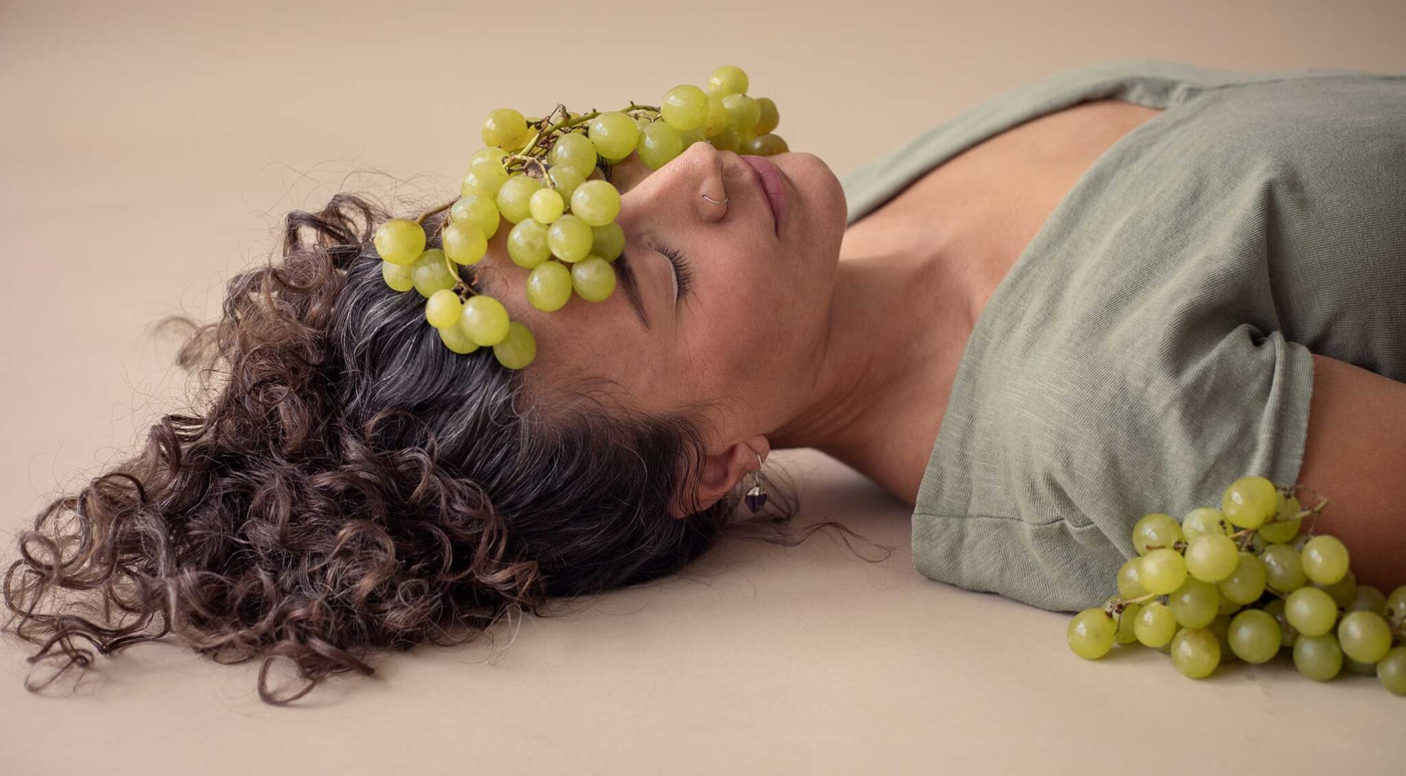 Laurie Ce., coach creativa, estirada y relajada. Medio rostro aparece cubierto por un racimo de uvas, con un toque orgánico y poético.